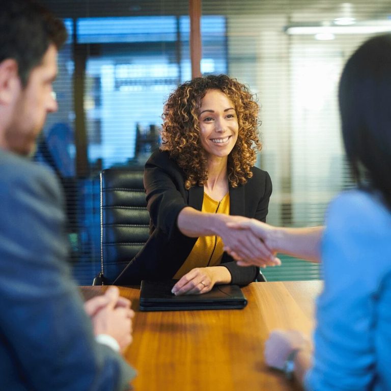 A woman and another woman shake hands in a professional office setting, symbolizing agreement or partnership.